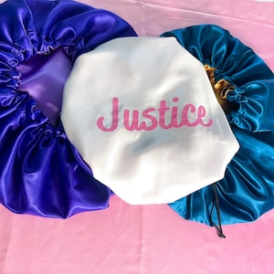 May include: Three satin hair bonnets, one white with the word "Justice" embroidered in pink, one teal blue, and one purple.