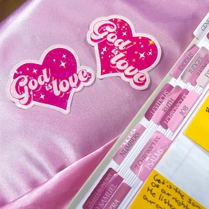 May include: Two pink heart-shaped stickers with a glittery finish. The stickers say "God is love" in a pink and white font with stars around the text.