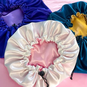 K&ouml;nnte beinhalten: Drei Hauben aus Satin mit verstellbaren Kordelz&uuml;gen und schwarzen Stoppern. Eine Haube ist hellrosa mit wei&szlig;em Rand, eine tiefblau mit violettem Innenfutter und eine dunkelblaugr&uuml;n mit goldenem Innenfutter, alle mit gerafftem Design.