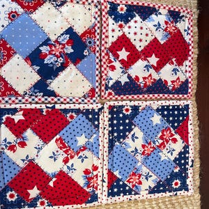 May include: Four square quilted coasters with a patriotic theme. Each coaster features a patchwork design with red, white, and blue fabrics. The patterns include floral prints, stars, and polka dots, with a red and white gingham border.
