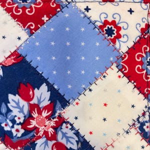 May include: Close-up of a patchwork textile with a patriotic theme. The design features red, white, and blue squares with floral and star patterns. The edges are stitched with red thread, and the border has a red, white, and blue checkered pattern.