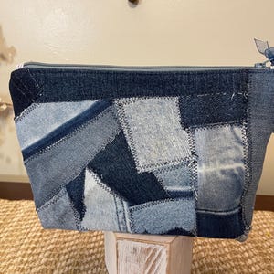 May include: A denim patchwork zippered pouch in various shades of blue. The rectangular bag features a patchwork design with visible stitching. A blue tassel and ribbon accent the zipper pull. The bag is sitting on a white wooden stand.