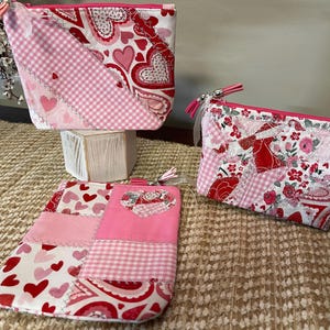 May include: Three handmade cosmetic bags in various sizes, featuring patchwork designs with pink, red, and white heart and floral patterns. Each bag has a zipper closure and a tassel. The bags are displayed on a woven surface.