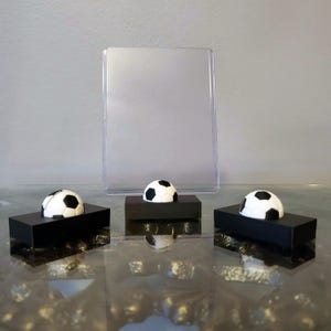 May include: Three miniature soccer balls on black bases with a clear acrylic sign holder in the background.