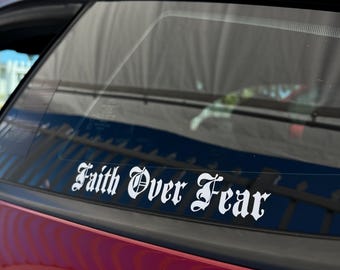 May include: A white vinyl decal with the phrase "Faith Over Fear" in an Old English font is affixed to the window of a red car. The decal is positioned horizontally.