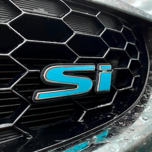 May include: A close-up of a black car grille with a blue "SI" emblem.