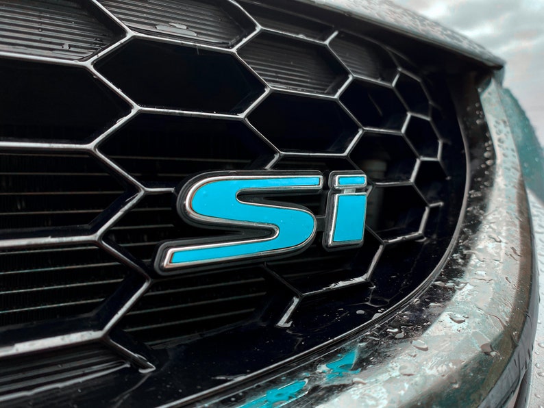 May include: A close-up of a black car grille with a blue "SI" emblem.