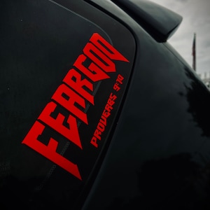 Puede incluir: Pegatina de vinilo roja con las palabras "FEAR GOD" en una fuente en negrita y estilizada. La pegatina está colocada en la ventana trasera de un coche negro. Debajo de las palabras, se lee "PROVERBS 9:10".