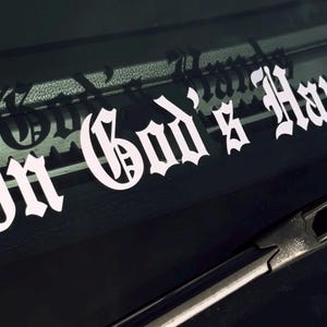 Può includere:  Adesivo in vinile bianco con il testo "In God's Hands" in uno stile di carattere gotico. L'adesivo è applicato sul parabrezza di un'auto nera.