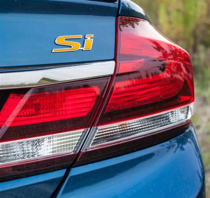 May include: A blue car with a red taillight and a yellow "Si" emblem on the trunk.