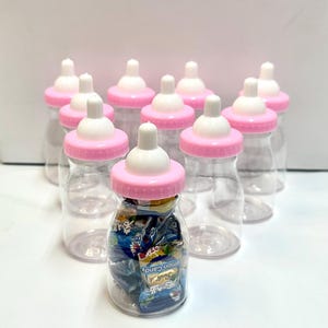 Puede incluir: Biberones de plástico transparente con tapas rosas y tetinas blancas, llenos de caramelos envueltos. Los biberones están dispuestos sobre una superficie blanca, lo que sugiere un tema de baby shower o fiesta. Los caramelos son visibles a través de los biberones transparentes.