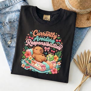 May include: Black t-shirt with a colorful graphic of a capybara relaxing on a pool float, with the text "Currently Avoiding Responsibilities". The design includes flowers, a frog, and cherries.