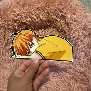 May include: A sticker featuring an anime character with orange hair and a yellow shirt. The character is depicted with their head resting on their arm. The sticker is held by a hand against a pink, fluffy background.