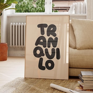 May include: A framed art print with the word "TRANQUILO" in black, bold letters. The print is leaning against a beige sofa in a bright room with a large window. A potted plant and wooden chair are also visible.