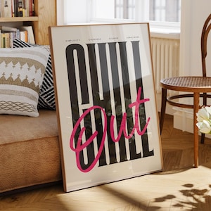 May include: A framed poster with the words "Chill Out" in black and pink, leaning against a sofa. The poster is in a room with a wooden floor, a chair, and a bookshelf filled with books. A vase of white flowers sits nearby.