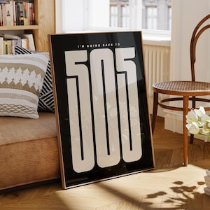 May include: A framed black and white art print with the text "I'M GOING BACK TO 505" in a bold, modern font. The print is displayed in a living room setting, leaning against a wall. The room features a bookshelf, a chair, and a vase of white flowers.