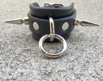 Puede incluir: Brazalete de muñeca de cuero negro con dos pinchos plateados y un anillo plateado. El brazalete tiene dos tachuelas plateadas.