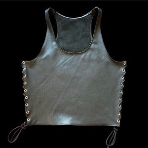Puede incluir: Un top de tirantes de cuero negro con cuello redondo y lados con cordones. Los cordones son de cordón negro y tienen ojales dorados.