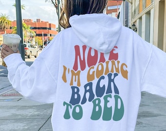 May include: White hoodie with the text "Nope I'm Going Back to Bed" in colorful retro lettering. The hoodie is worn by a person walking on a sidewalk. The person is wearing white sneakers and socks. The background includes buildings and a street.