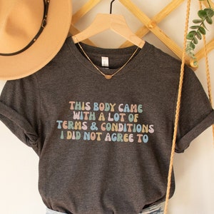 May include: A dark gray t-shirt with the text "This body came with a lot of terms & conditions I did not agree to" in rainbow colors. The shirt is hanging on a wooden hanger with a straw hat and a woven bag.