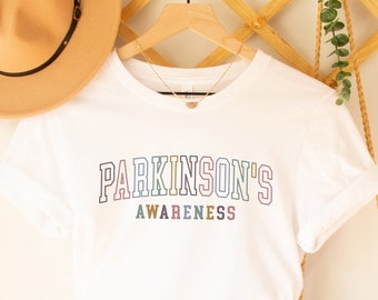 May include: White t-shirt with the text "PARKINSON'S AWARENESS" in rainbow colors. The shirt is hanging on a wooden hanger with a straw hat and a woven bag.