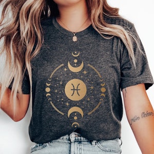May include: A dark grey t-shirt with a gold graphic of the Pisces zodiac sign surrounded by moons and stars. The text "Pisces" is written on the shirt.
