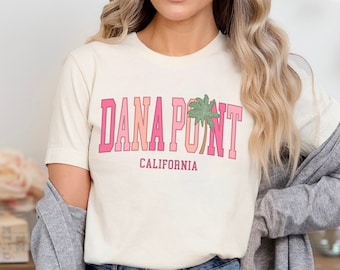 Puede incluir: Una mujer lleva una camiseta blanca con un gráfico rosa y rojo que dice "Dana Point, California" y una palmera. También lleva pantalones cortos de mezclilla y una chaqueta de punto gris.