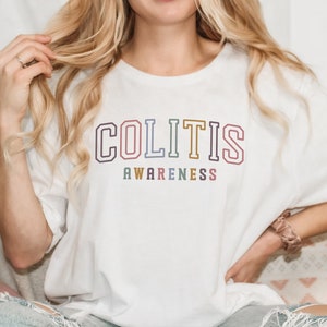 May include: White t-shirt with the words "COLITIS AWARENESS" in a colorful, collegiate-style font. The shirt is a casual, unisex style, perfect for raising awareness. The design features a rainbow color scheme.