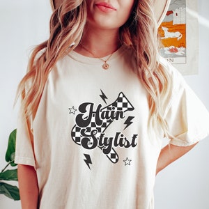 May include: A white t-shirt with a black and white checkered design featuring the text "Hair Stylist" in a retro font. The design includes lightning bolts and stars.