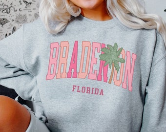 Puede incluir: Una sudadera gris con un gráfico rosa y naranja que dice "Bradenton, Florida" con un diseño de palmera.