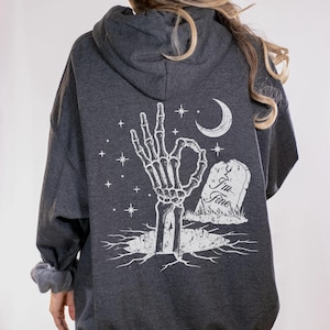 May include: Dark gray hoodie with a white graphic on the back. The graphic features a skeleton hand making an "okay" gesture, a crescent moon, stars, and a tombstone with the text "I'm Fine."