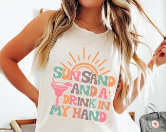 Puede incluir: Camiseta blanca de tirantes con un gráfico colorido que dice "Sun, Sand and a Drink in My Hand". El gráfico presenta un sol, una copa de cóctel y un arcoíris de colores.