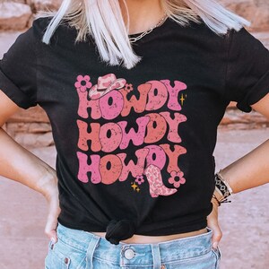 May include: Black t-shirt featuring a pink and red graphic design that says "Howdy" three times. The graphic also includes a cowboy hat and boot.