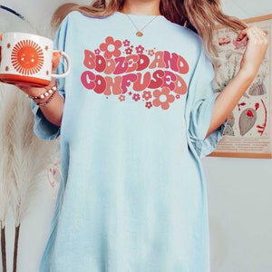 May include: An oversized light blue t-shirt featuring a retro-style graphic that reads "Boozed and Confused" in pink and red lettering with pink and red flowers surrounding the text.
