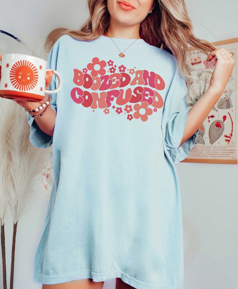 May include: An oversized light blue t-shirt featuring a retro-style graphic that reads "Boozed and Confused" in pink and red lettering with pink and red flowers surrounding the text.
