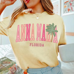 May include: A yellow sweatshirt with the text "ANNA MARIA FLORIDA" in pink and coral lettering, with a palm tree graphic. The sweatshirt is a casual style.