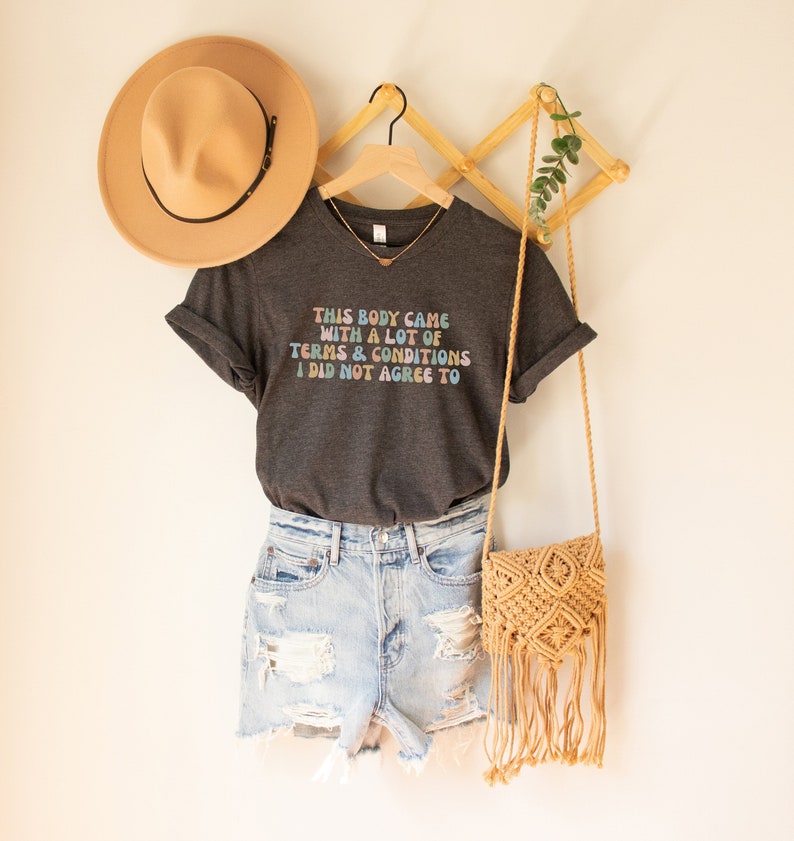 May include: A dark gray t-shirt with the text "This body came with a lot of terms & conditions I did not agree to" in rainbow colors. The shirt is hanging on a wooden hanger with a straw hat and a woven bag.