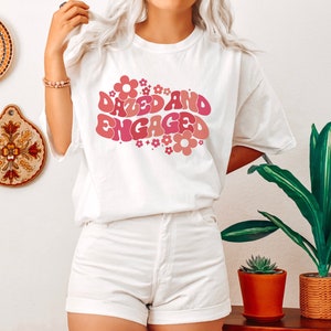 May include: White t-shirt with a pink and red graphic that says "Dazed and Engaged" with flowers around the text.