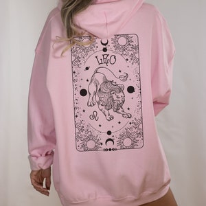 May include: A light pink hoodie with a black graphic on the back. The graphic features a lion, the word "LEO", suns, moons, and stars. The hoodie has a hood and long sleeves.