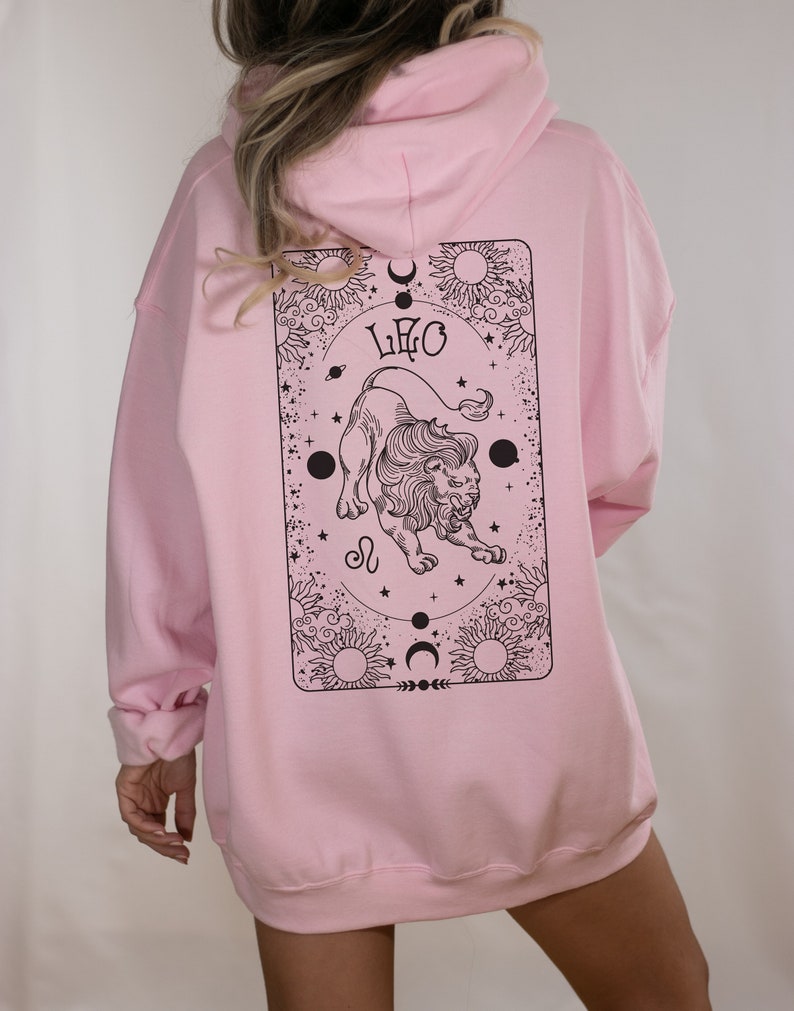 May include: A light pink hoodie with a black graphic on the back. The graphic features a lion, the word "LEO", suns, moons, and stars. The hoodie has a hood and long sleeves.