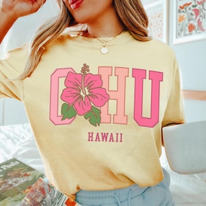 May include: Yellow sweatshirt with the word "OHU" in pink block letters, a pink hibiscus flower, and the word "HAWAII" in a matching font. The sweatshirt is a light yellow color.