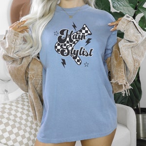 May include: A light blue t-shirt with a black and white checkered hairdryer graphic and the text "Hair Stylist" in a retro font.
