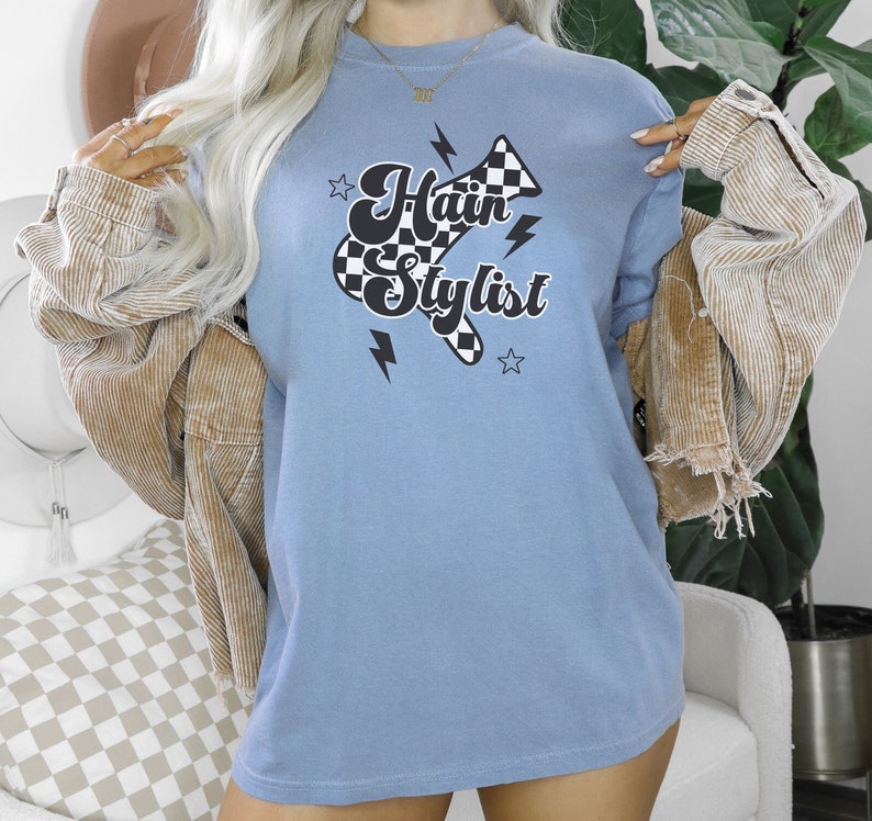 May include: A light blue t-shirt with a black and white checkered hairdryer graphic and the text "Hair Stylist" in a retro font.