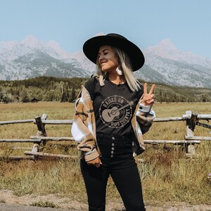 May include: A person wearing a black hat, black t-shirt with a guitar graphic, and a plaid shirt. The t-shirt says "Raised on 90's Country". The background features a mountain range, a wooden fence, and a field.