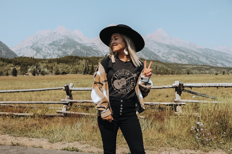 May include: A person wearing a black hat, black t-shirt with a guitar graphic, and a plaid shirt. The t-shirt says "Raised on 90's Country". The background features a mountain range, a wooden fence, and a field.