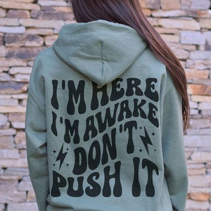 May include: A sage green hooded sweatshirt with the text "I'm here I'm awake don't push it" printed in black on the back.