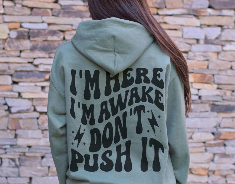 May include: A sage green hooded sweatshirt with the text "I'm here I'm awake don't push it" printed in black on the back.