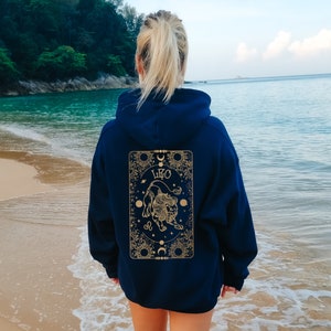 May include: Navy blue hoodie with a gold Leo zodiac design on the back. The design features a lion illustration, the word "LEO", and celestial elements. The hoodie is worn on a beach with the ocean in the background.