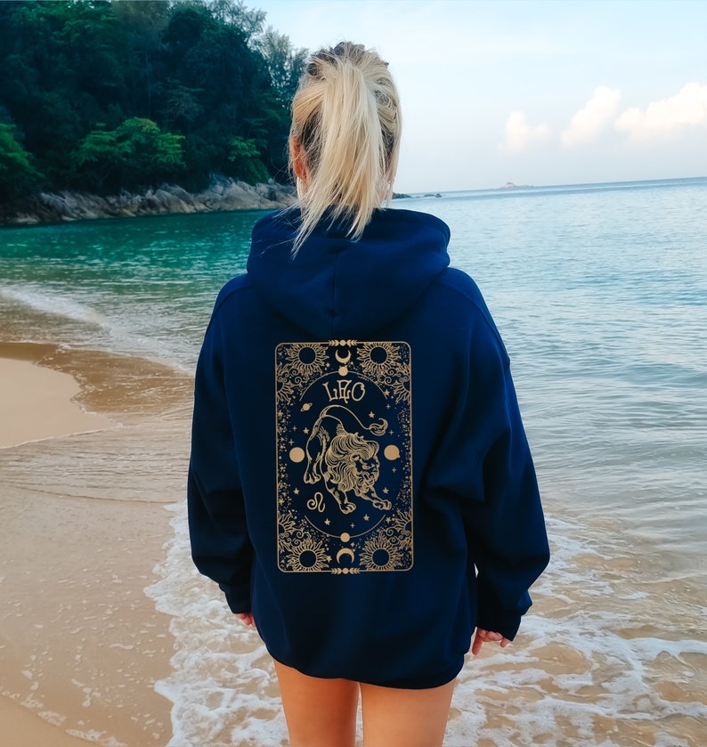 May include: Navy blue hoodie with a gold Leo zodiac design on the back. The design features a lion illustration, the word "LEO", and celestial elements. The hoodie is worn on a beach with the ocean in the background.
