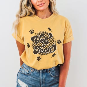 Puede incluir: Una camiseta amarilla mostaza con un gr&aacute;fico de estilo retro que dice "Vet Tech" en una fuente marr&oacute;n. El gr&aacute;fico est&aacute; rodeado de huellas de patas y rayos.